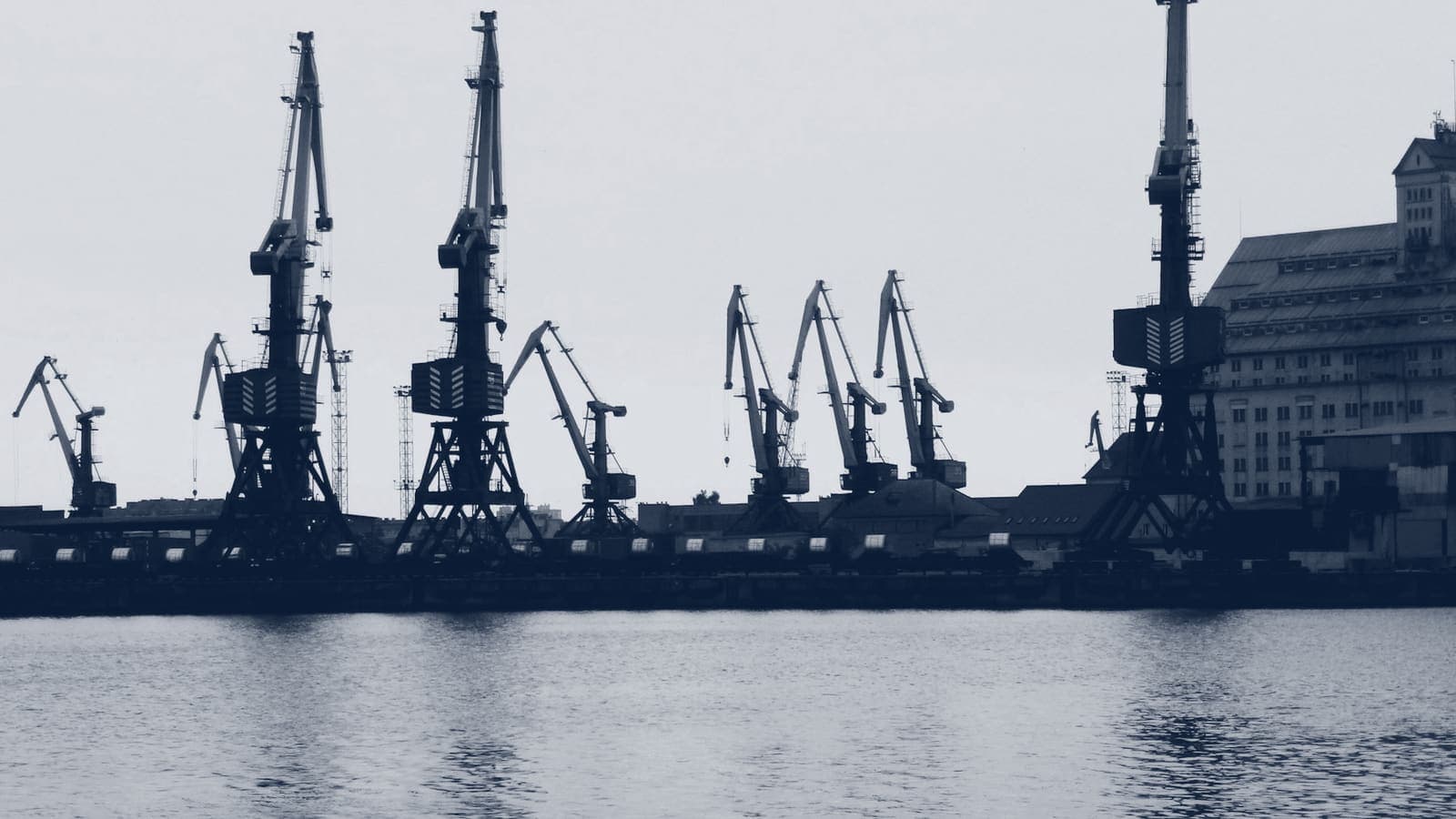 Row of harbour cranes in silhouette along a quay, seen across calm water