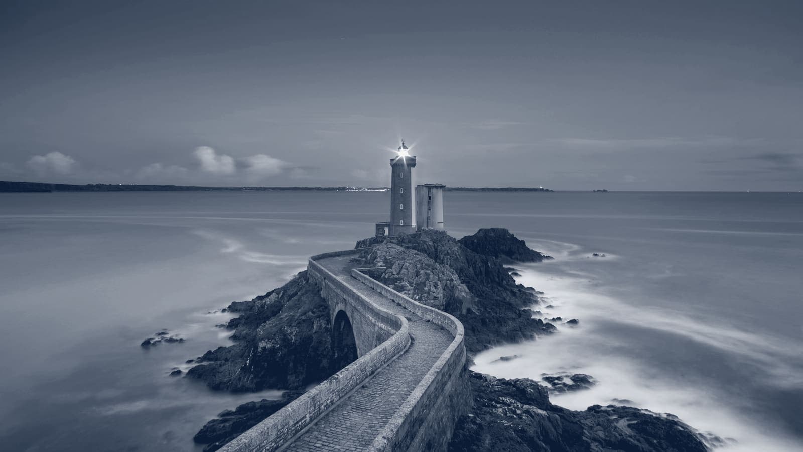 Stone causeway curving across rocks to a lit lighthouse on a small islet