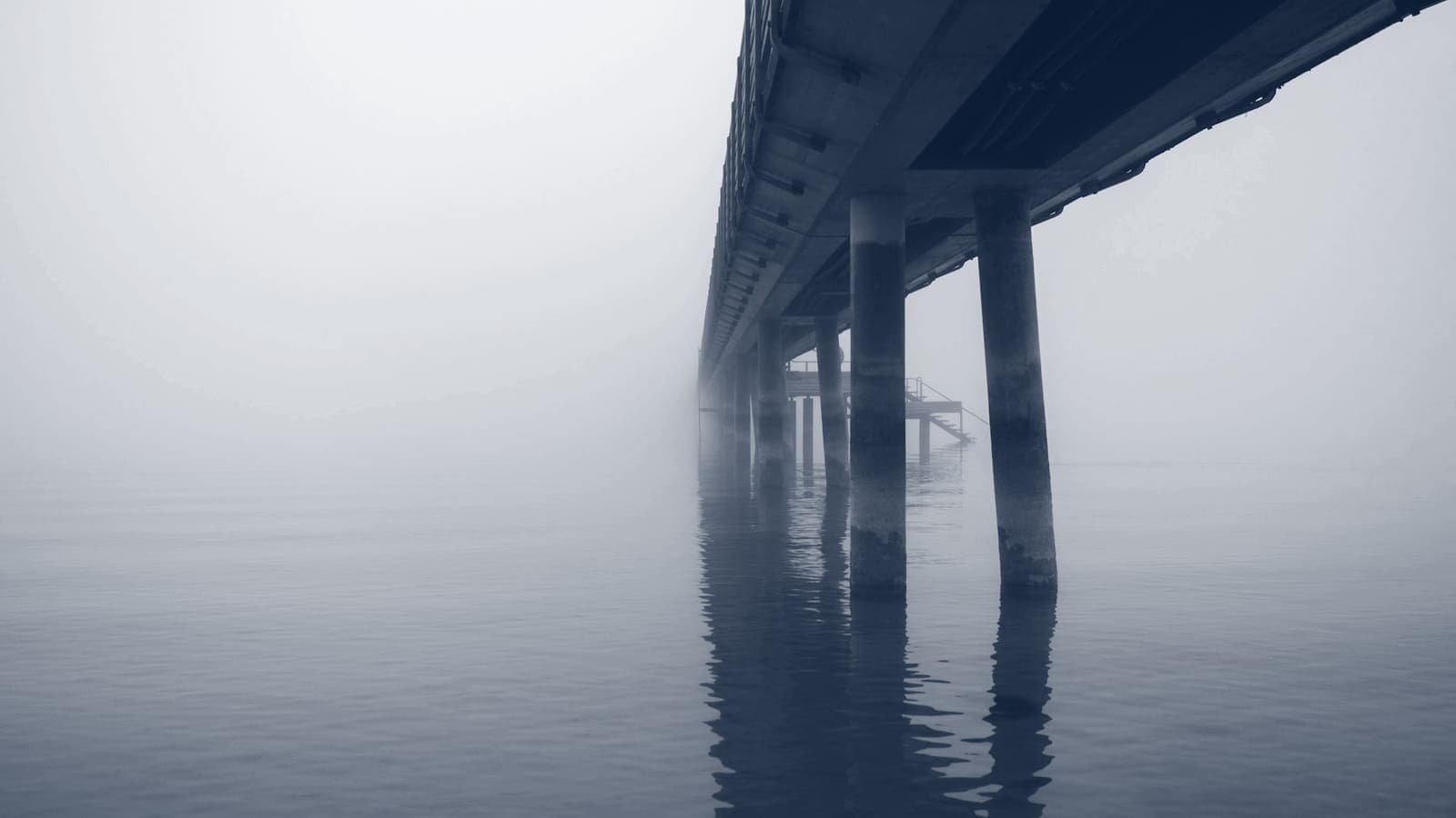 Underside of a long pier on pillars disappearing into thick fog over still water